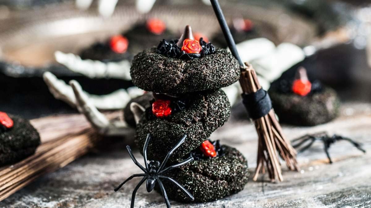 Three dark cookies decorated with red icing and chocolate pieces are stacked with a toy spider, broom, and skeleton hand nearby, evoking a Halloween theme.