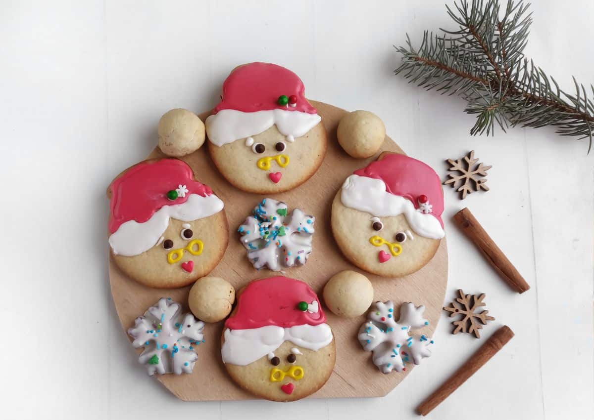 A round wooden board with decorated Christmas cookies shaped like Santa faces, snowflakes, and round cookies, next to cinnamon sticks, star anise, and a pine branch.
