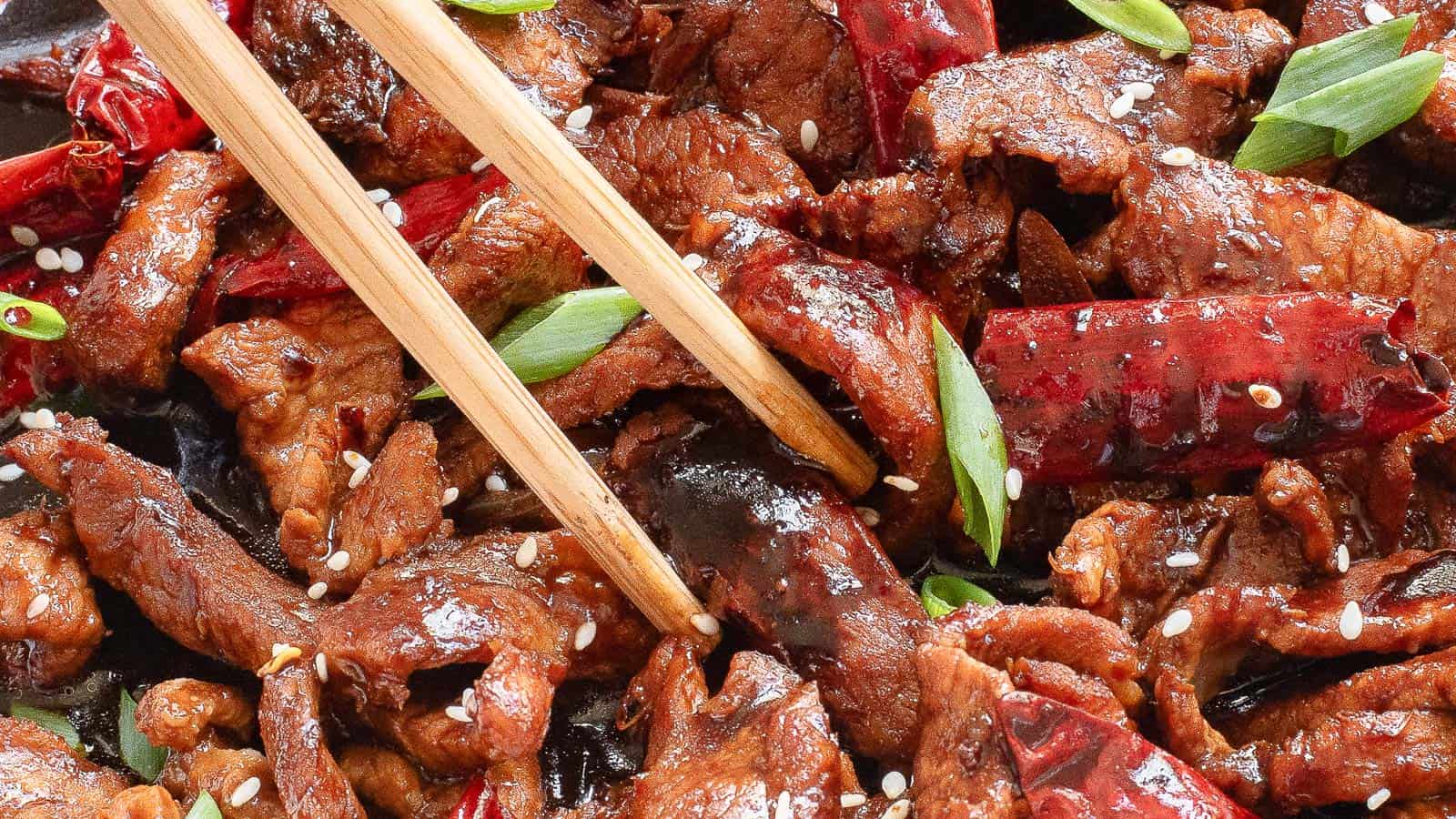 Close-up of a spicy beef stir-fry with dried red chilies, green onions, sesame seeds, and chopsticks.