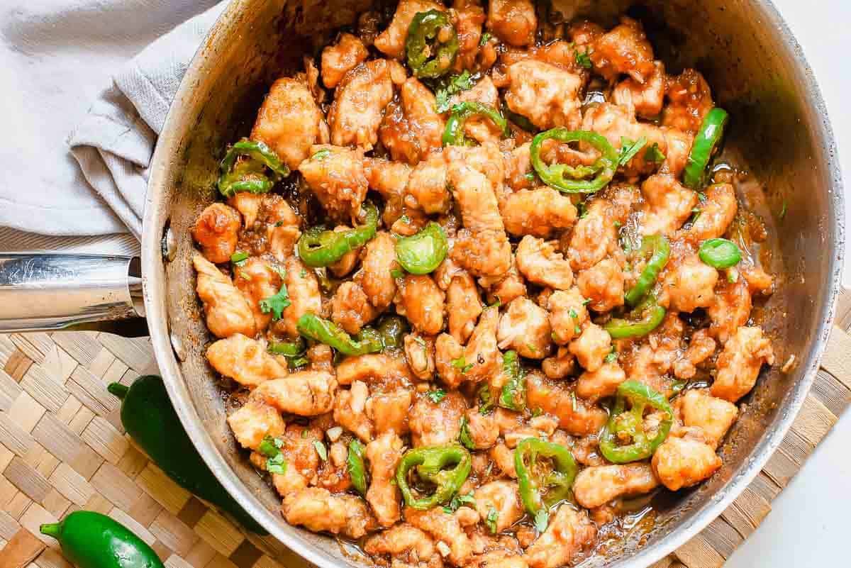 A skillet filled with cooked chicken pieces in a brown sauce, garnished with sliced green jalapeños, sits on a woven placemat.