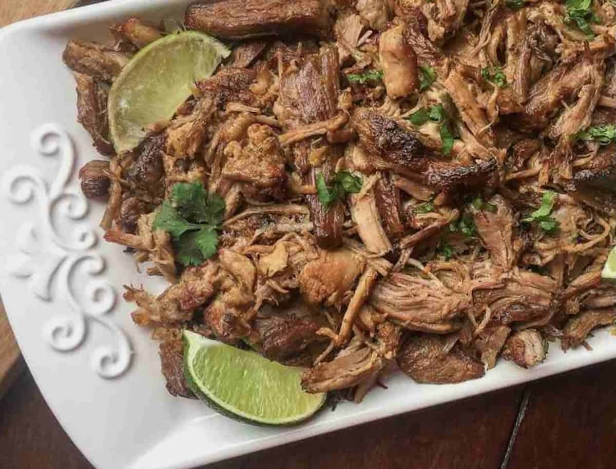 Shredded cooked meat garnished with cilantro and lime wedges, served on a white rectangular plate.