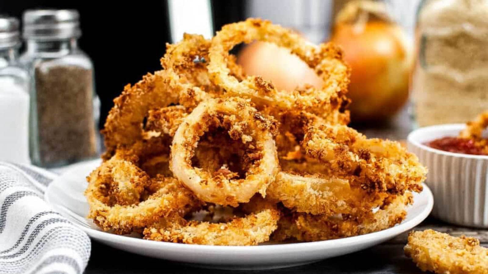 A plate of crispy golden onion rings sits on a table, with salt and pepper shakers, an onion, and a bowl of dipping sauce in the background.