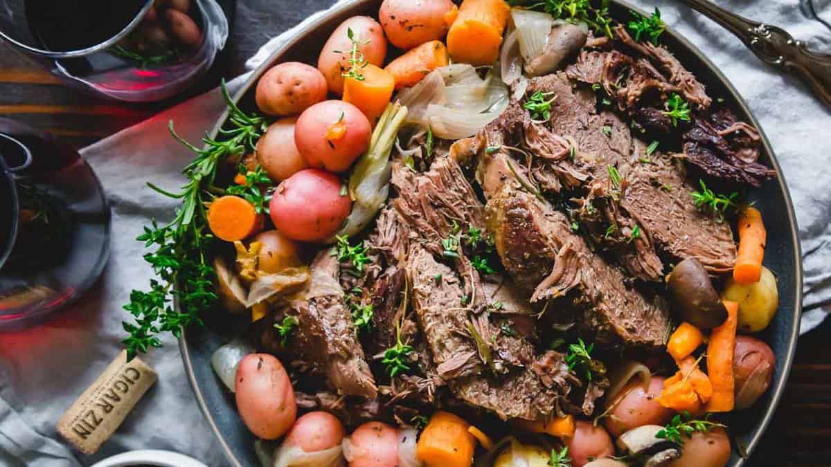 A platter of sliced pot roast surrounded by baby potatoes, carrots, onions, and garnished with fresh herbs.