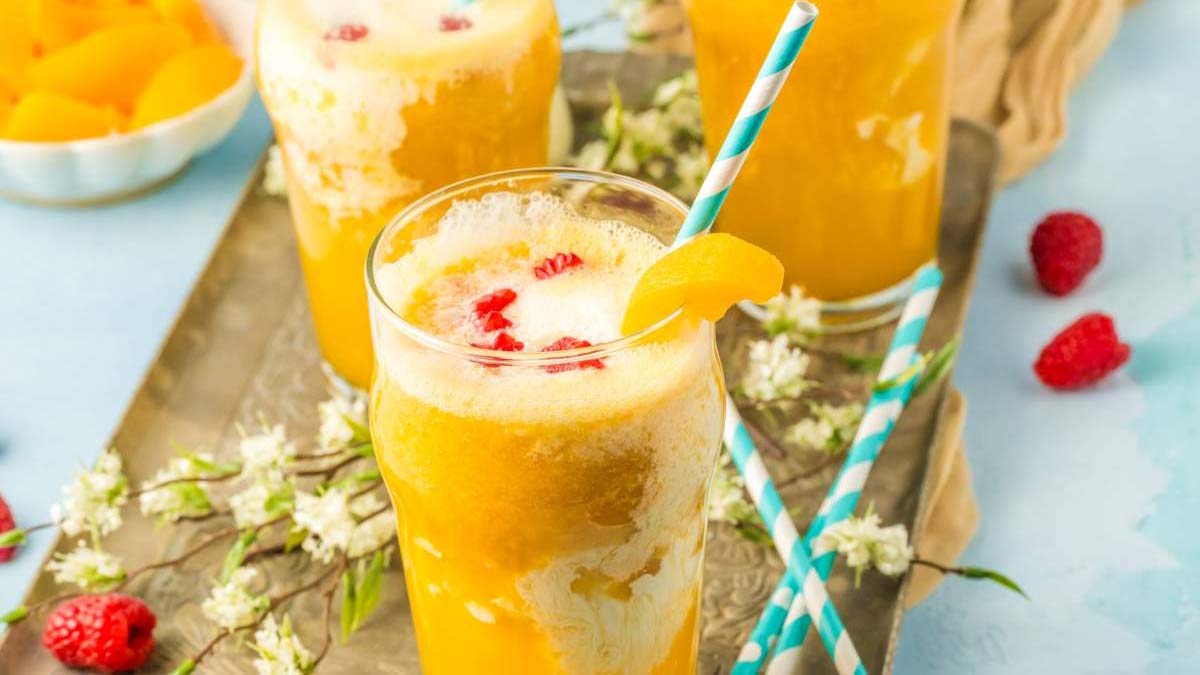 Three glasses of peach and orange smoothie with blue-striped straws, topped with peach slices and raspberries, on a tray with flowers and more raspberries.