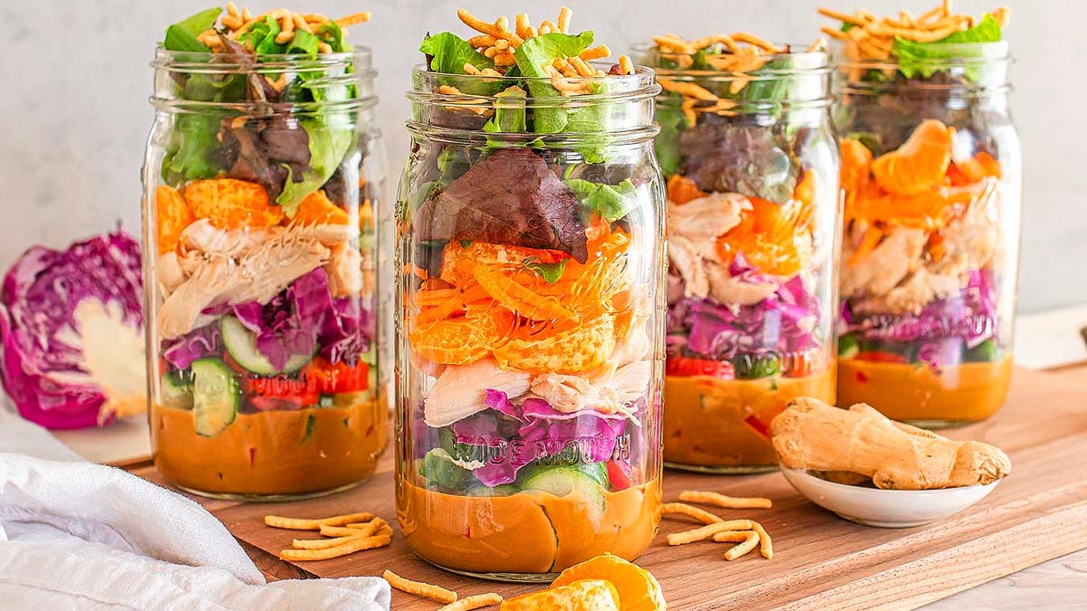 Four mason jars filled with layered salad ingredients including lettuce, chicken, mandarin slices, shredded carrots, cucumber, and purple cabbage, set on a wooden surface.
