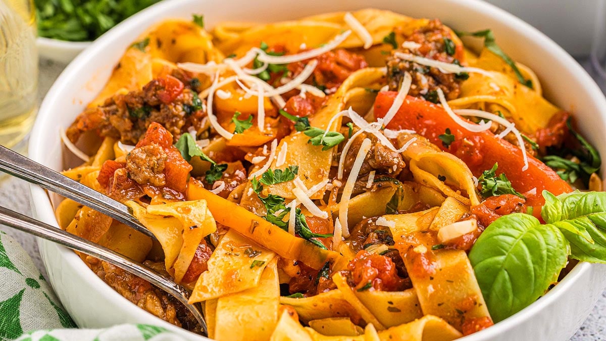 A bowl of pappardelle pasta with tomato sauce, ground meat, vegetables, and grated cheese, garnished with parsley and basil, served with a fork and spoon.