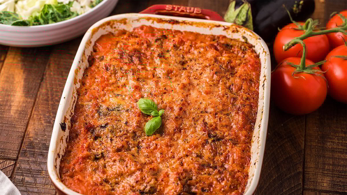 A baked dish of lasagna topped with melted cheese and herbs, garnished with basil, sits on a wooden table next to fresh tomatoes and an eggplant.