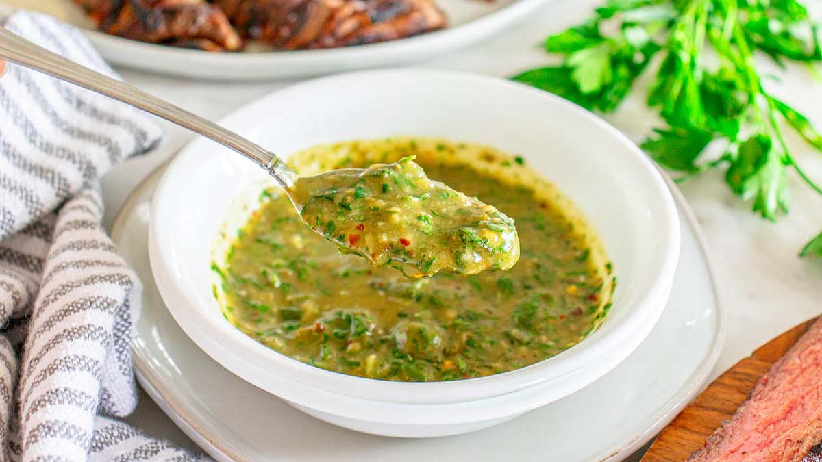 A spoon lifts a portion of green chimichurri sauce from a white bowl, with fresh parsley and sliced meat visible in the background.
