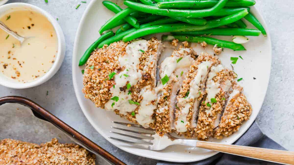 A plate with sliced, breaded chicken topped with creamy sauce, green beans on the side, and a fork; extra sauce in a small bowl nearby.