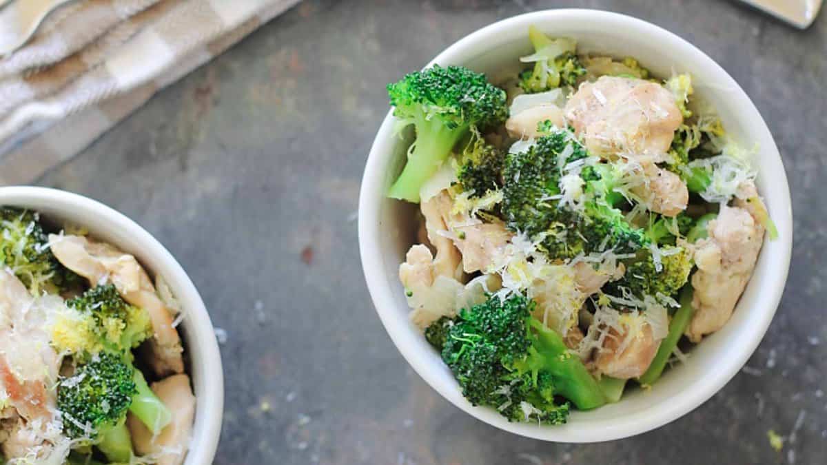 Two white bowls filled with a mixture of cooked chicken, broccoli florets, and grated cheese, placed on a dark surface.
