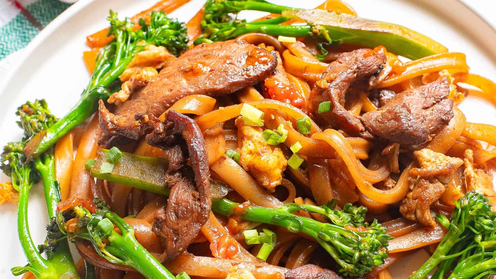 A plate of stir-fried rice noodles with beef, broccolini, eggs, and scallions in a brown sauce, served on a white dish.