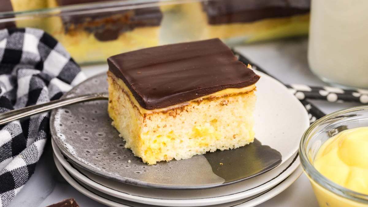 A slice of yellow cake with a chocolate frosting layer is served on a gray plate with a fork beside it.