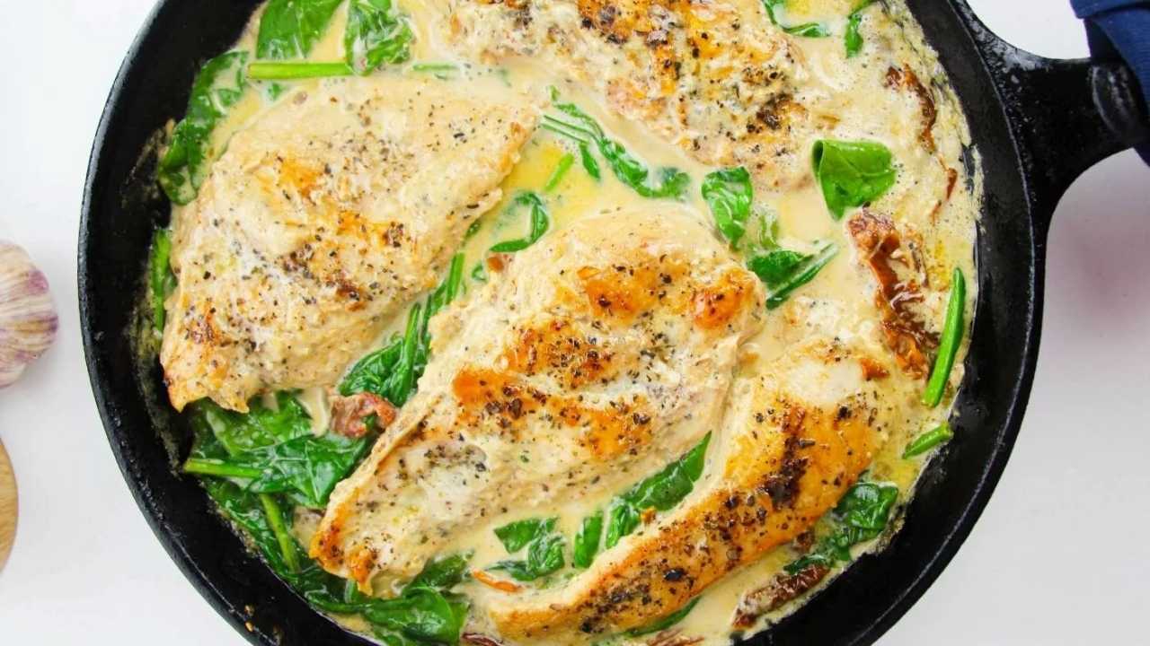 Three seasoned chicken breasts in a creamy sauce with spinach and sun-dried tomatoes, cooked in a black skillet.