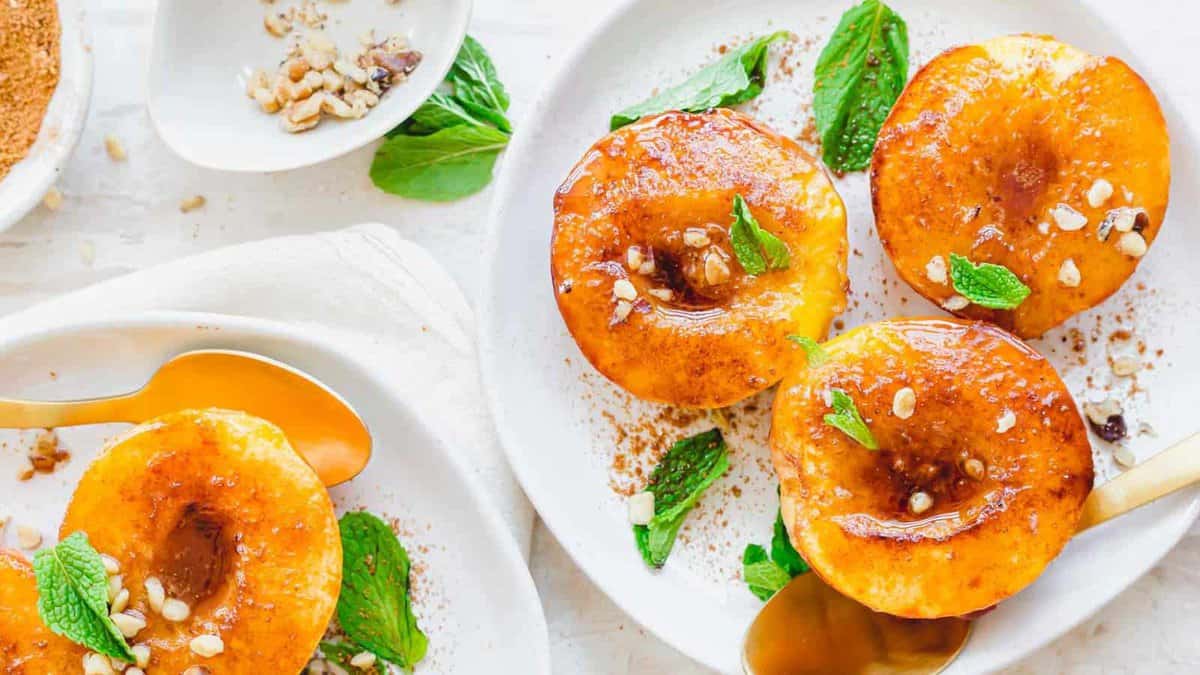 Three baked peaches topped with caramel sauce, mint leaves, and chopped nuts on a white plate, with a gold spoon and extra toppings on the side.