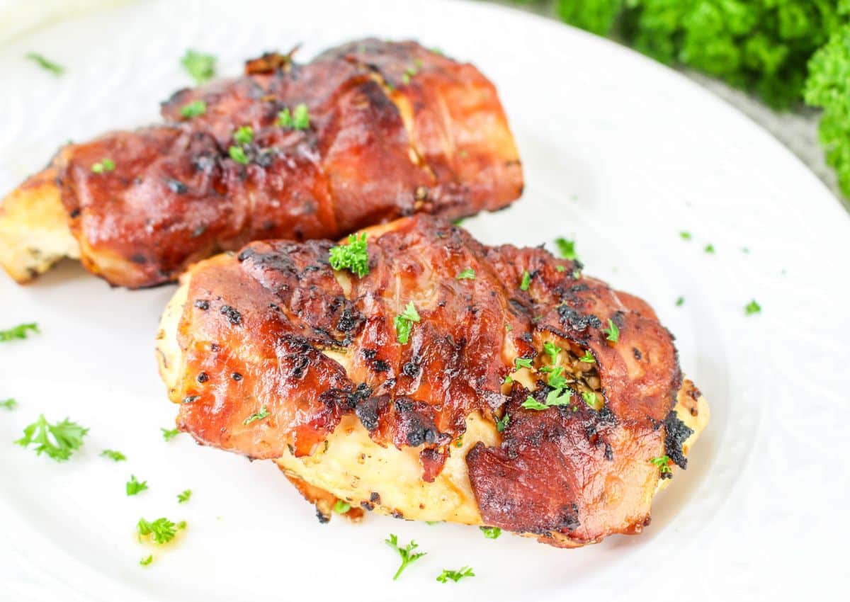 Two pieces of cooked chicken breast wrapped in crispy bacon, garnished with chopped parsley, served on a white plate.