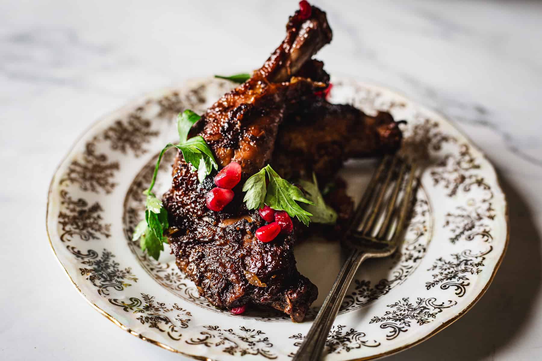 Two pieces of cooked, seasoned chicken garnished with fresh parsley and pomegranate seeds on a patterned plate with a fork.