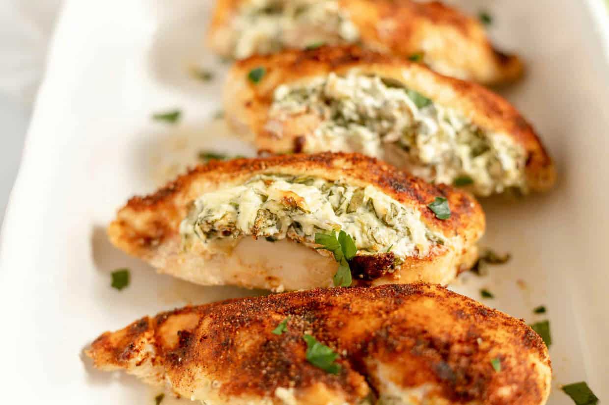 Four seasoned chicken breasts sliced open and stuffed with a creamy mixture of herbs and cheese, arranged on a white serving dish and garnished with chopped parsley.