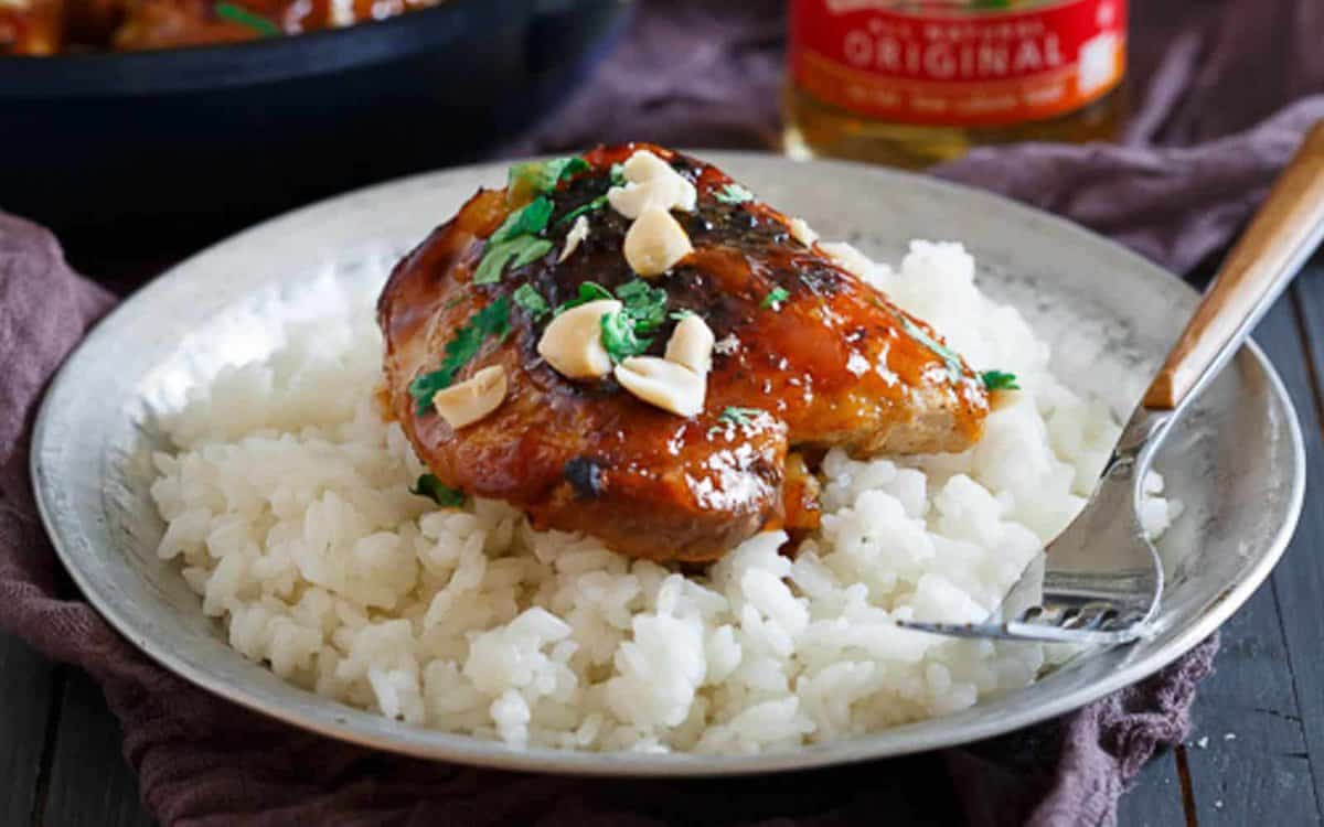 A plate of white rice topped with glazed chicken, garnished with chopped nuts and herbs, with a fork on the side.