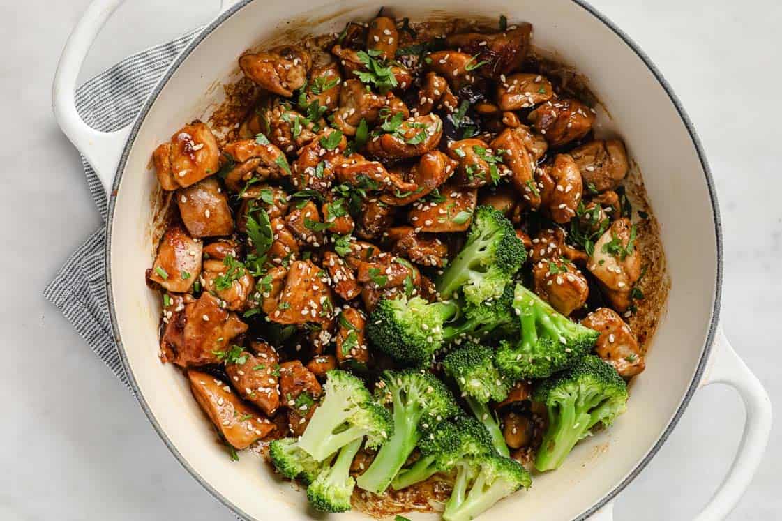 Cooked chicken pieces with sauce, topped with sesame seeds and chopped herbs, are served in a white pot alongside steamed broccoli florets.