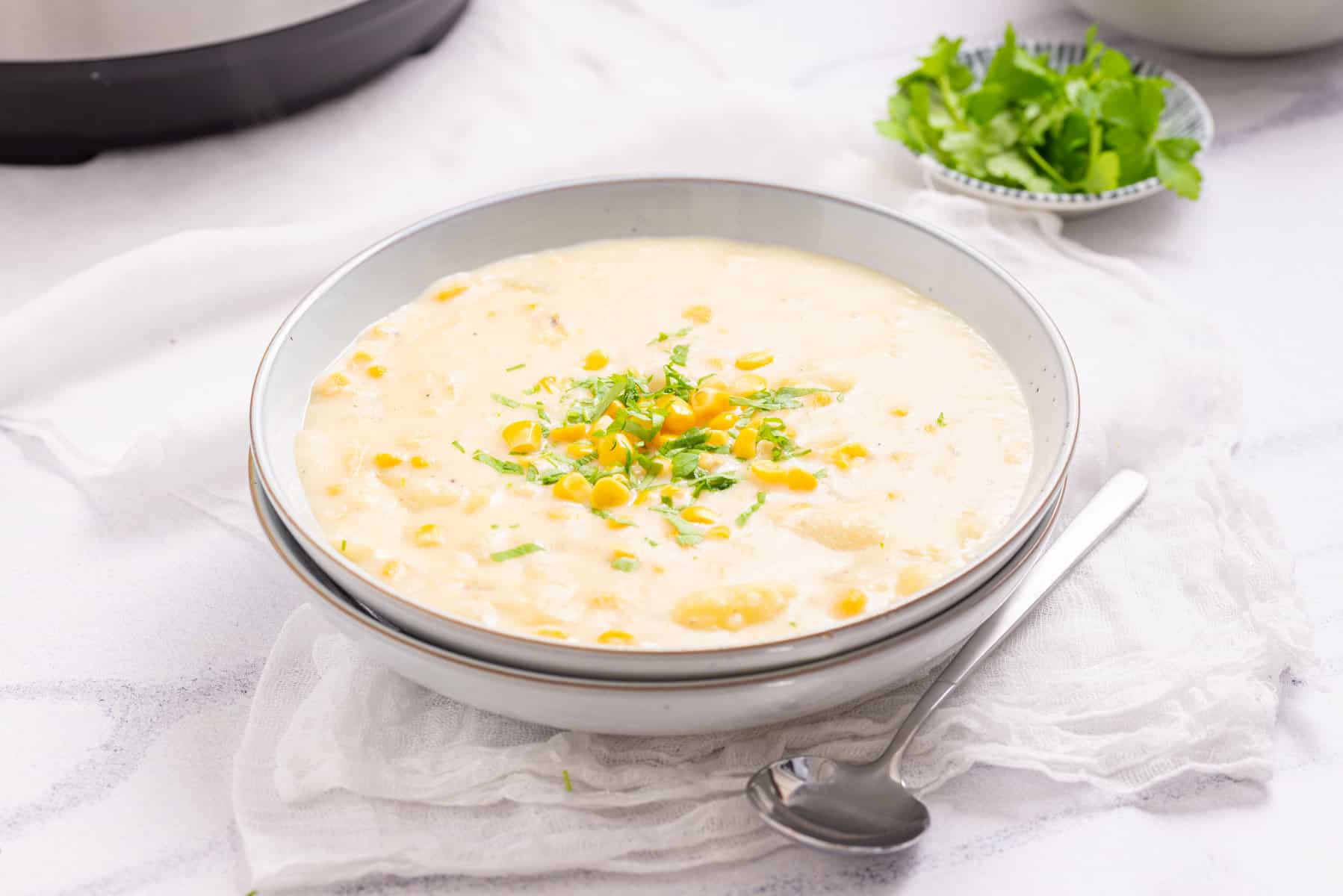 A bowl of creamy corn chowder garnished with chopped green herbs, placed on a white cloth next to a spoon and a small dish of fresh herbs.