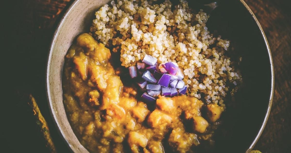 A bowl containing cooked quinoa, yellow curry with vegetable chunks, and a garnish of chopped purple onions.