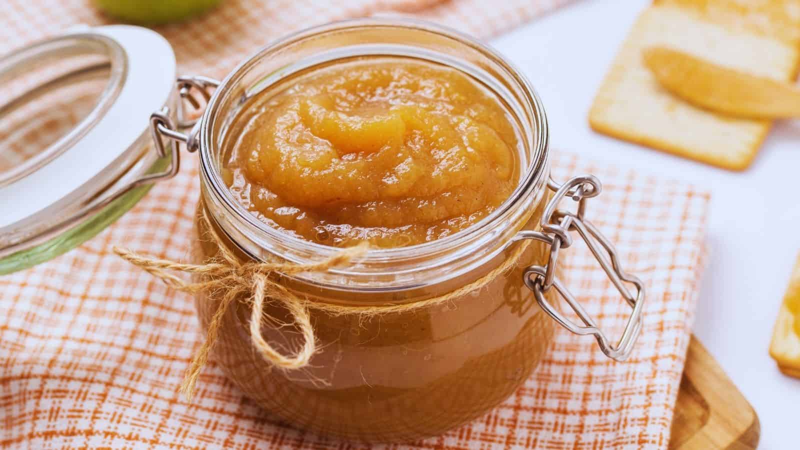 An open glass jar filled with applesauce sits on a checkered cloth, with crackers and more applesauce in the background.