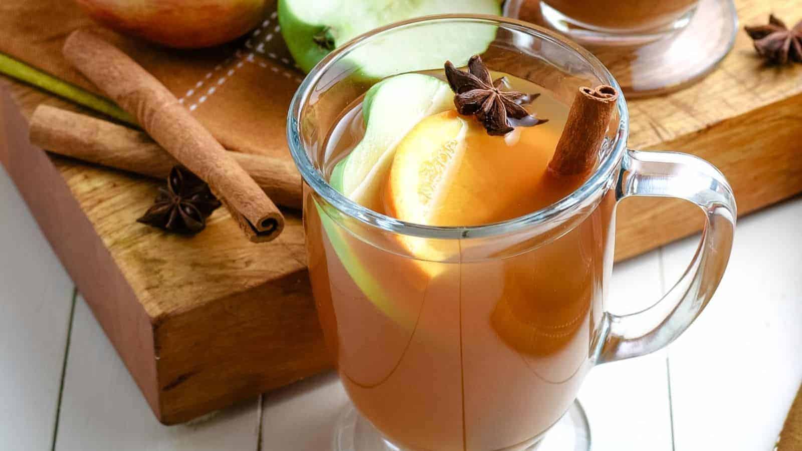 A glass mug of spiced apple cider with apple slices, a cinnamon stick, and star anise, placed on a wooden board with whole cinnamon sticks and a green apple.