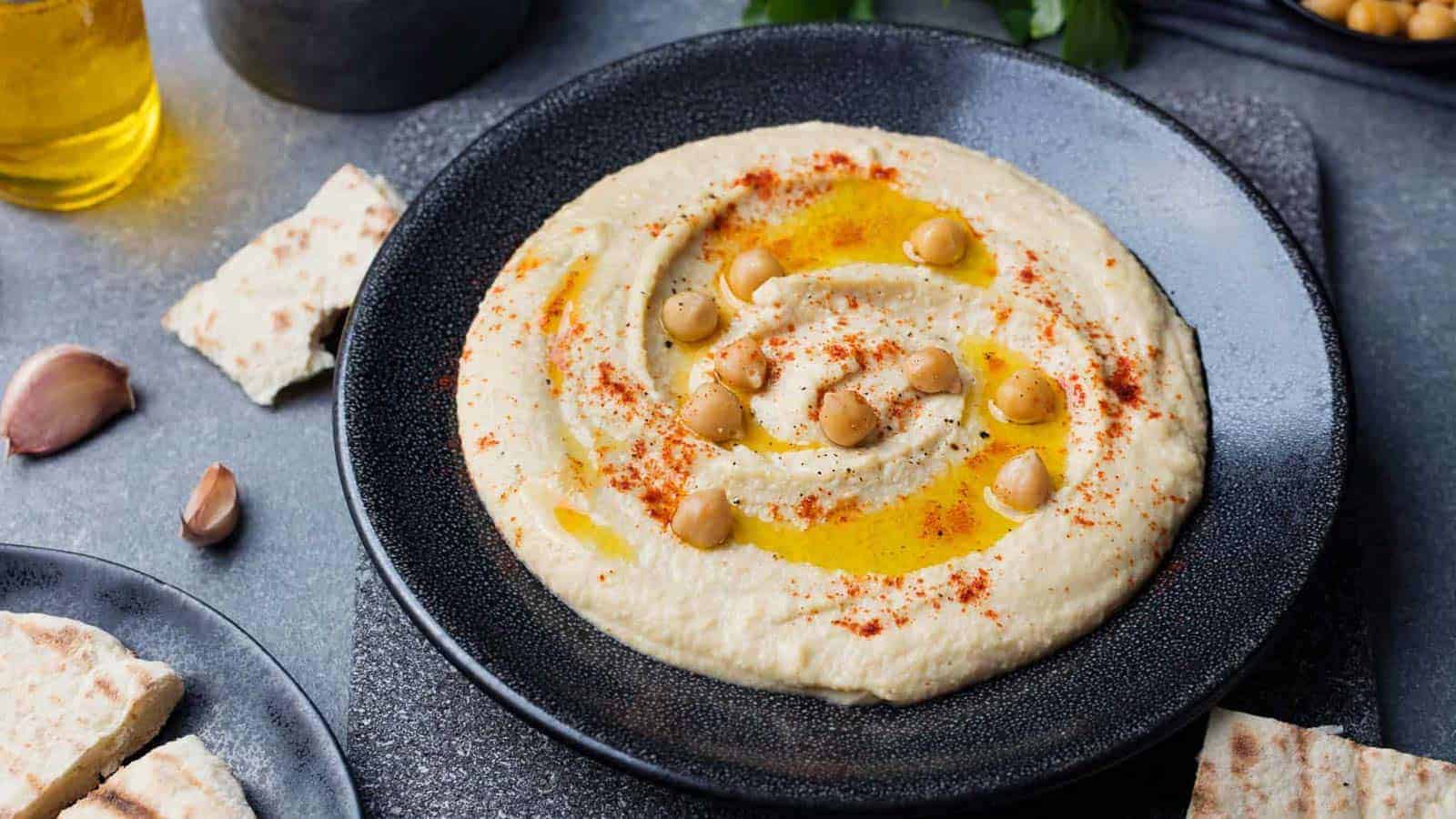 A bowl of hummus topped with whole chickpeas, paprika, and olive oil, served with pita bread on the side.