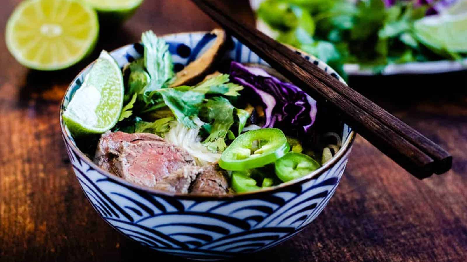 A bowl of noodle soup topped with sliced beef, jalapeños, lime, cilantro, cabbage, and mushrooms, with chopsticks resting on the bowl.