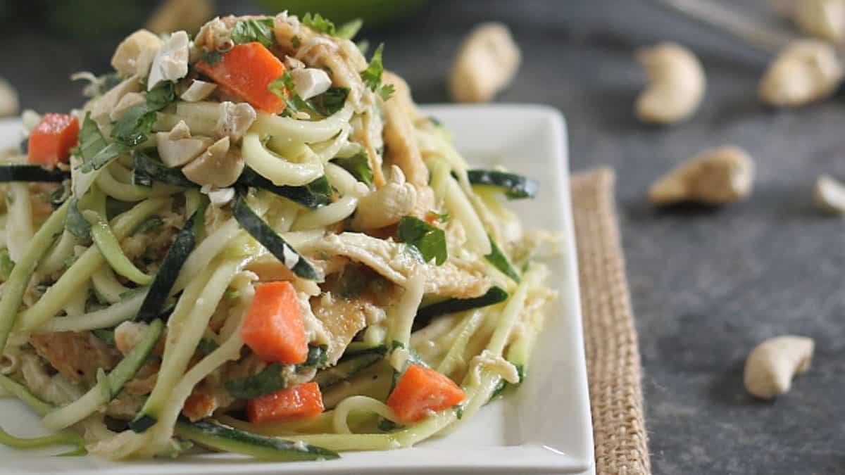 A plate of zucchini noodles topped with chopped carrots, herbs, and cashew nuts sits on a white square dish.