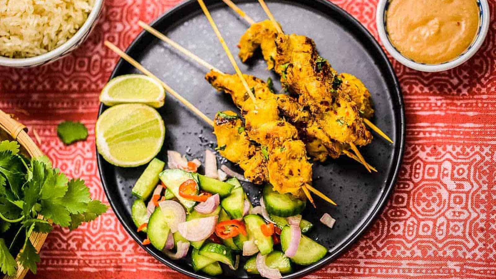 Grilled chicken skewers on a black plate with sliced lime, cucumber salad, and a bowl of peanut sauce on a red patterned tablecloth.