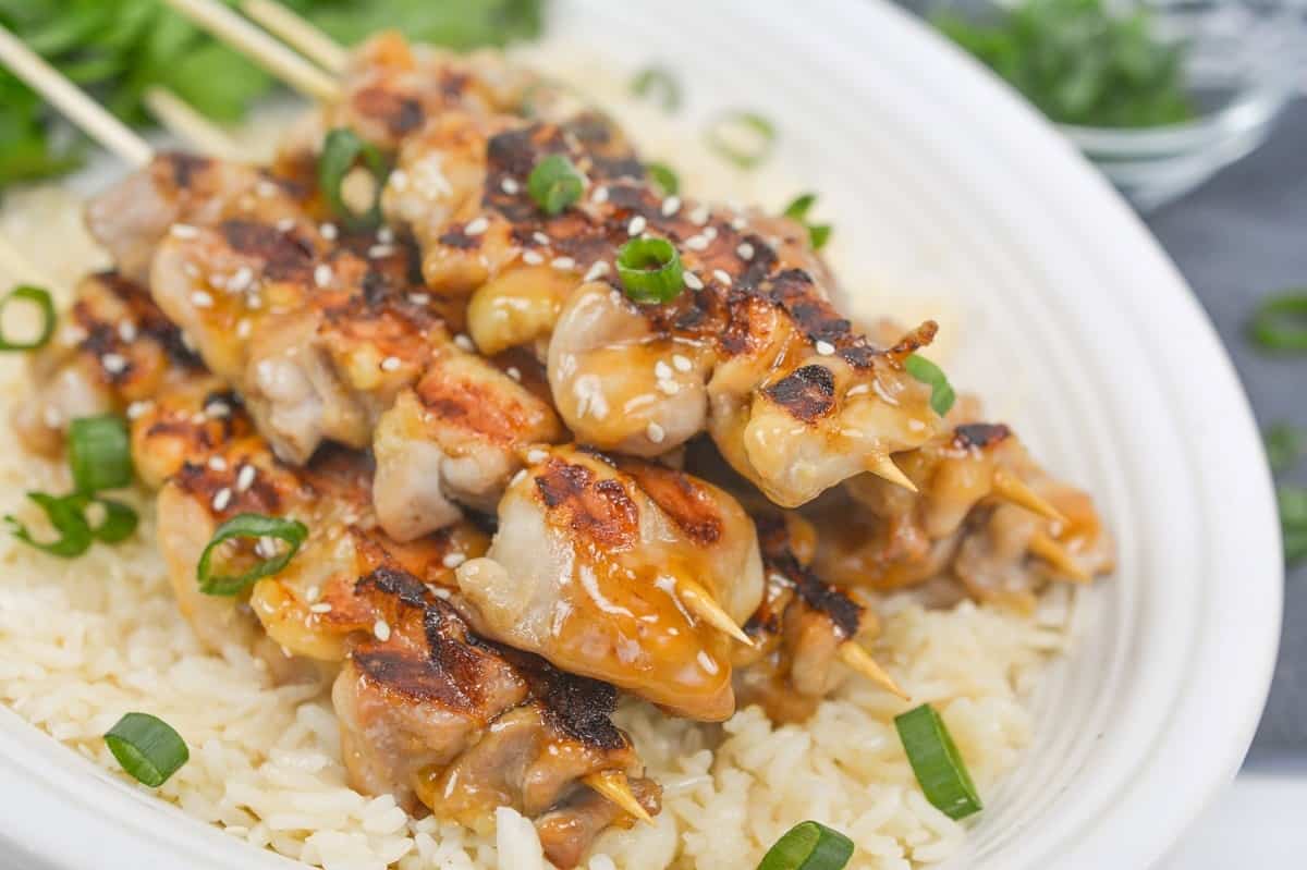 Grilled chicken skewers with sesame seeds and sliced green onions served over white rice on a white plate.