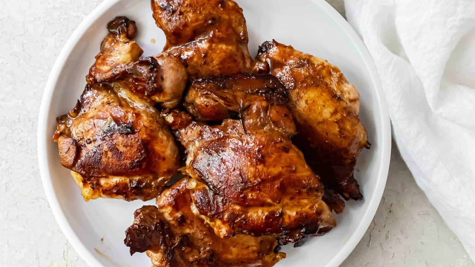 Plate with several pieces of cooked, glazed chicken thighs on a white surface, next to a white napkin.