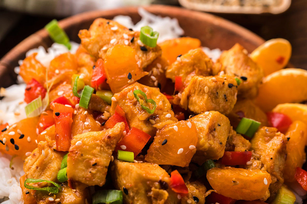 A close-up of a bowl filled with white rice, topped with cooked chicken pieces, orange segments, red bell peppers, green onions, and sesame seeds.