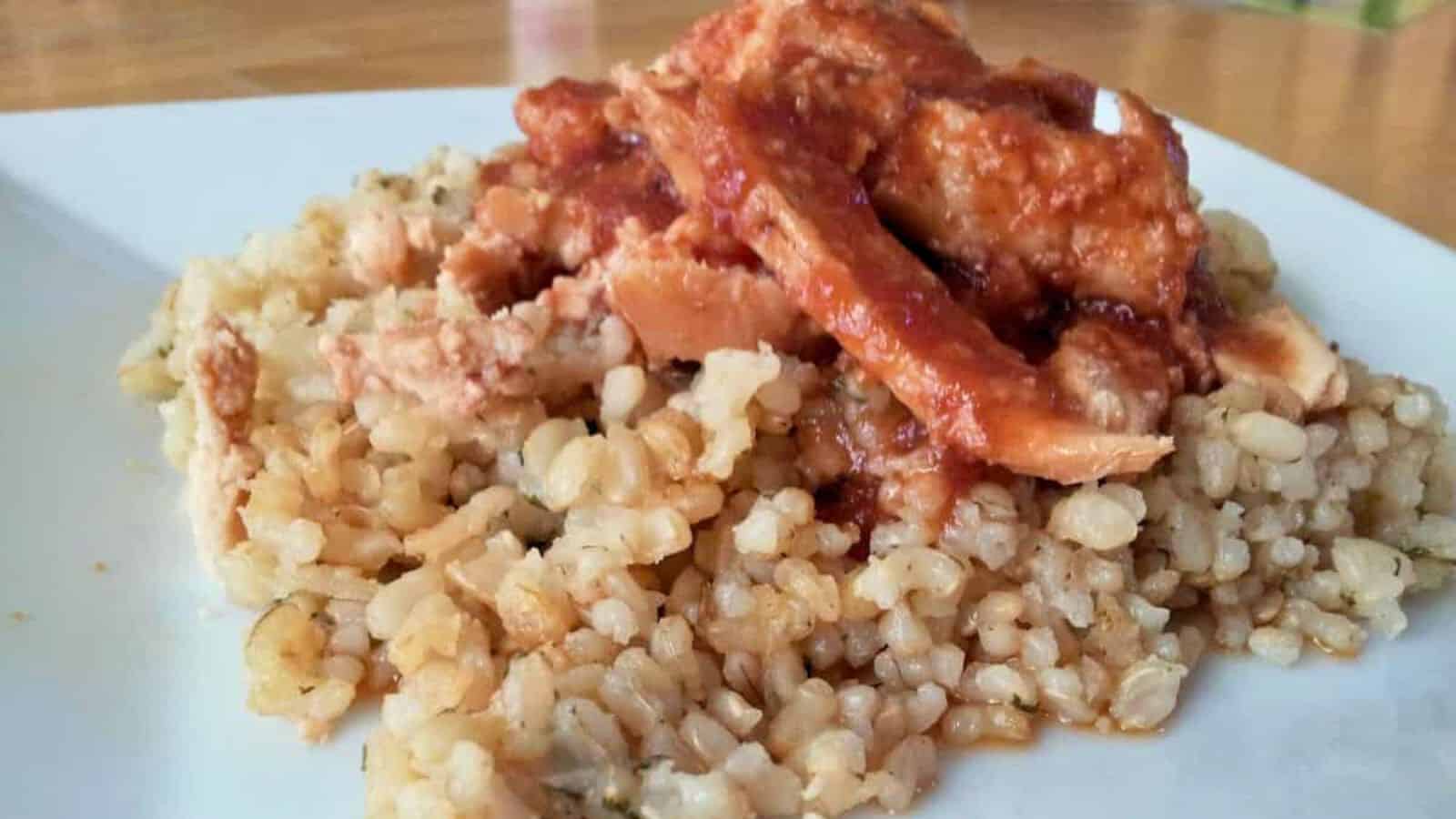 A serving of cooked brown rice topped with sliced chicken and tomato-based sauce on a white plate.