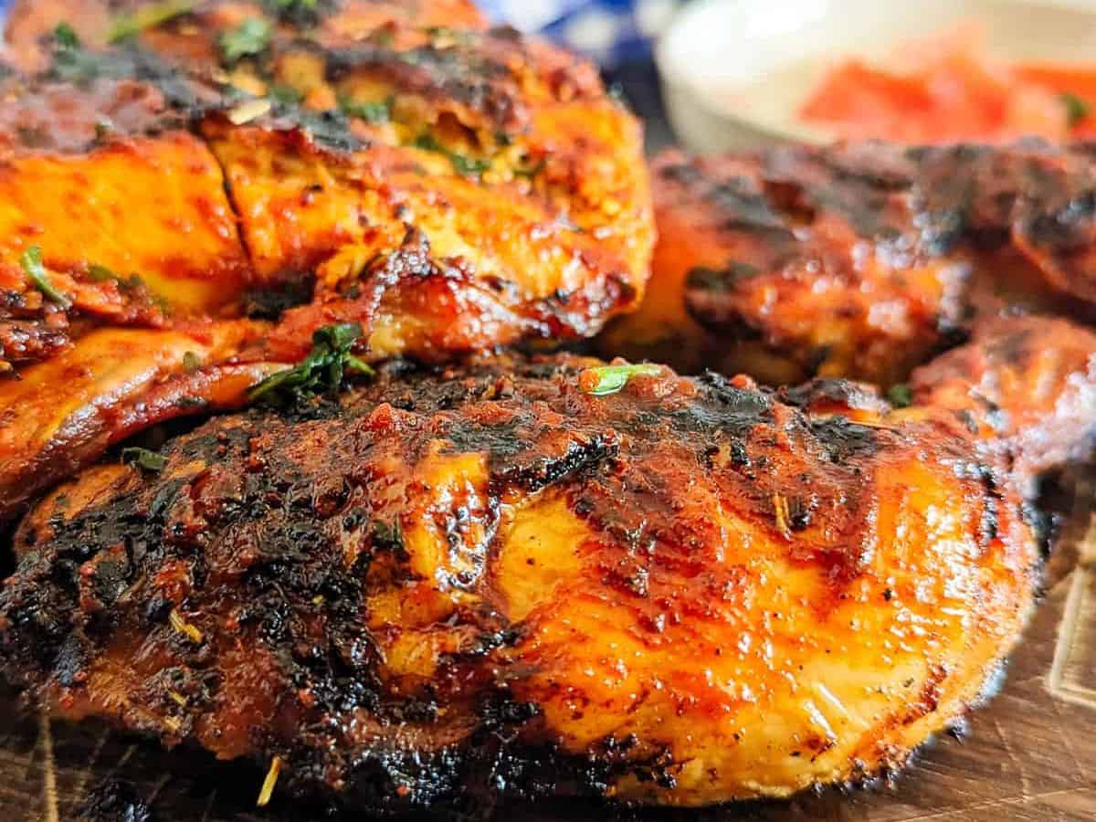 Close-up of grilled, seasoned chicken pieces with a charred surface, garnished with herbs and served on a wooden board.