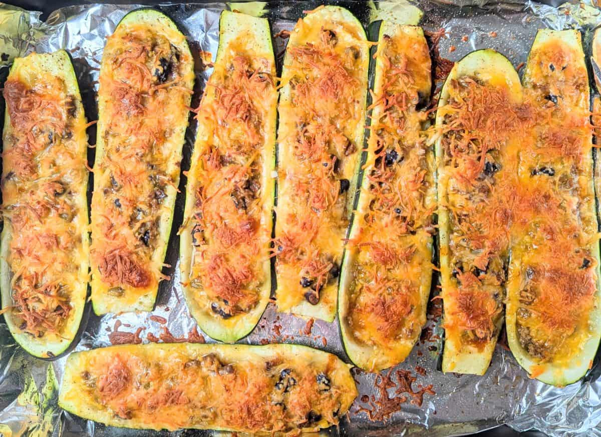 Eight zucchini halves topped with melted cheese and baked filling, arranged on a foil-lined baking sheet.