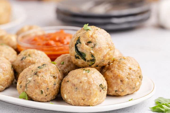 A plate of cooked turkey meatballs with herbs, stacked in the center, with a small bowl of marinara sauce in the background.