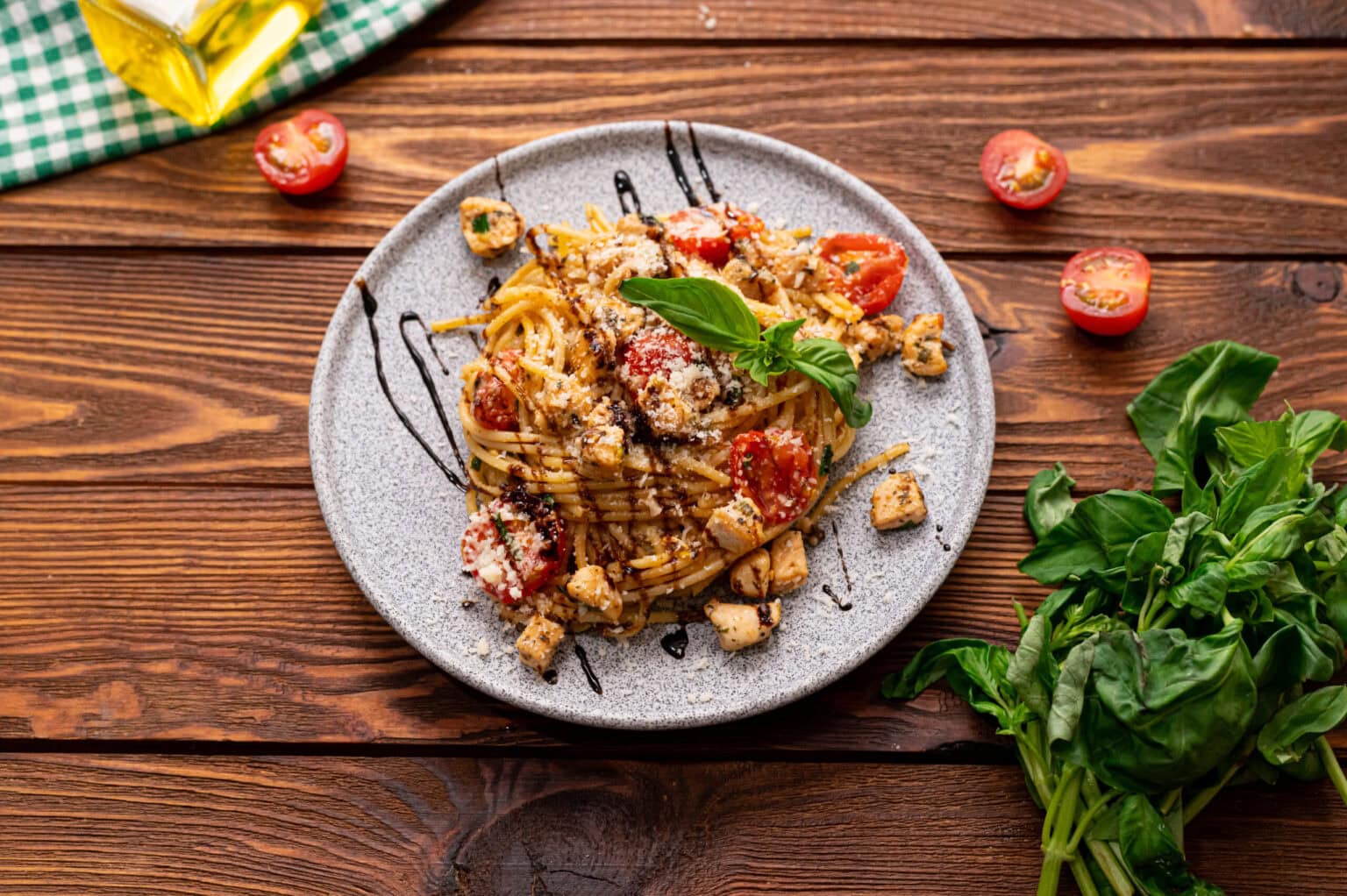 A plate of spaghetti with grilled chicken, cherry tomatoes, basil, grated cheese, and balsamic glaze on a wooden table, next to fresh basil and a green-checked cloth.