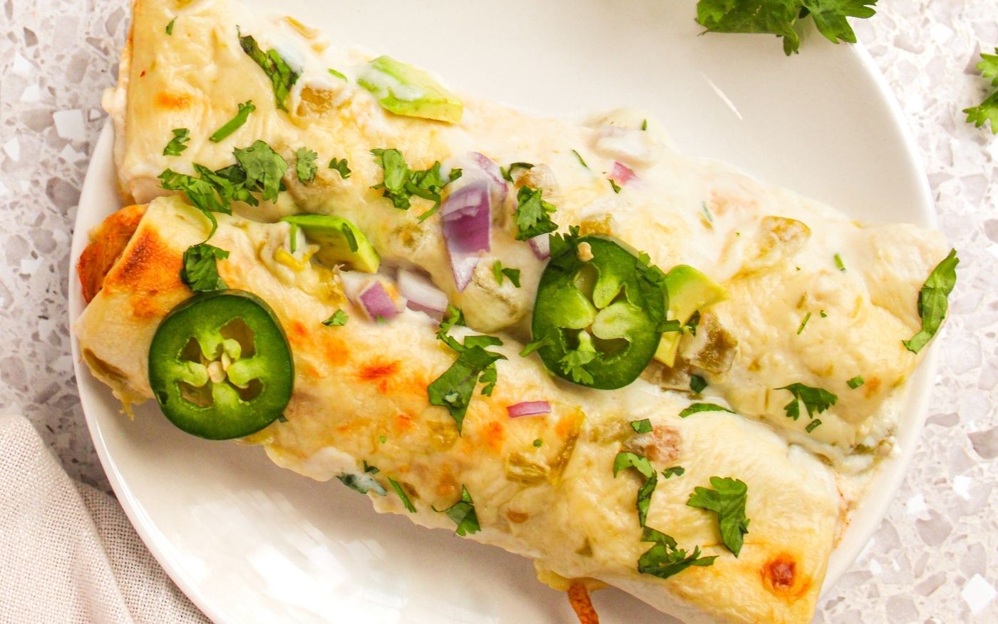 A cheesy enchilada topped with sliced jalapeños, chopped red onion, and fresh cilantro on a white plate.