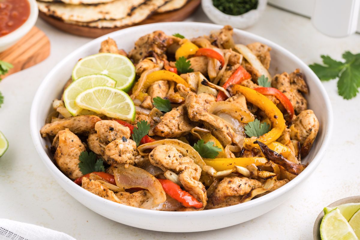 A bowl of chicken fajitas with grilled chicken strips, red and yellow bell peppers, onions, cilantro, and lime slices. Tortillas and salsa are visible in the background.