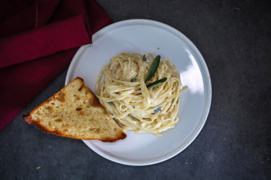 A plate of creamy fettuccine pasta garnished with herbs, served with a slice of toasted bread on the side, placed on a dark surface next to a folded red napkin.