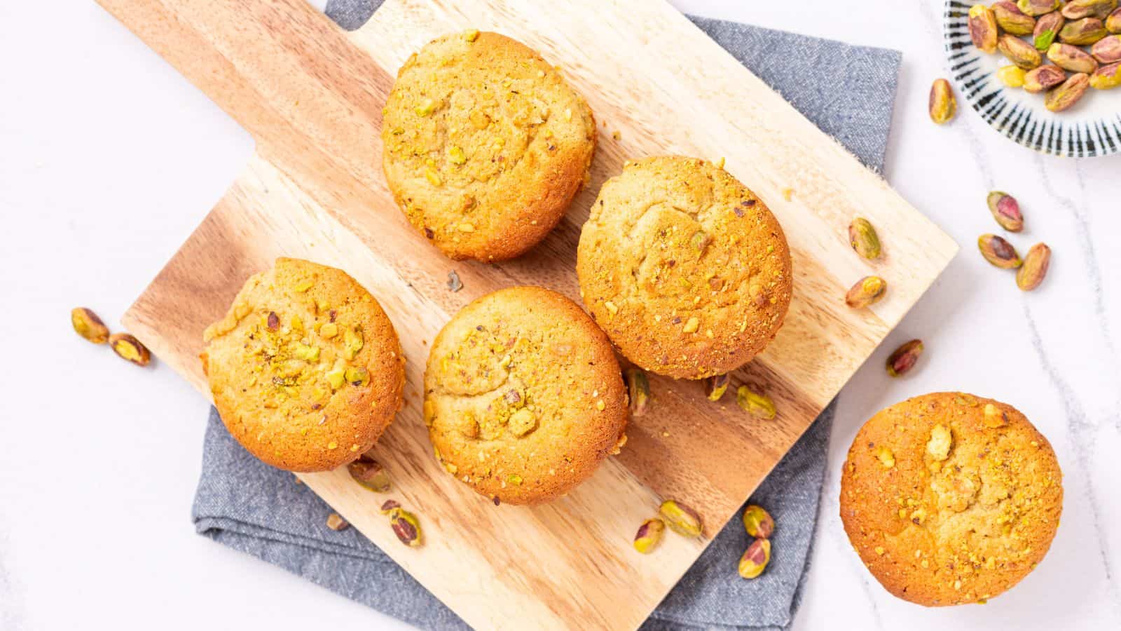 Five pistachio muffins are arranged on a wooden board with a gray cloth underneath, with whole pistachios scattered around.