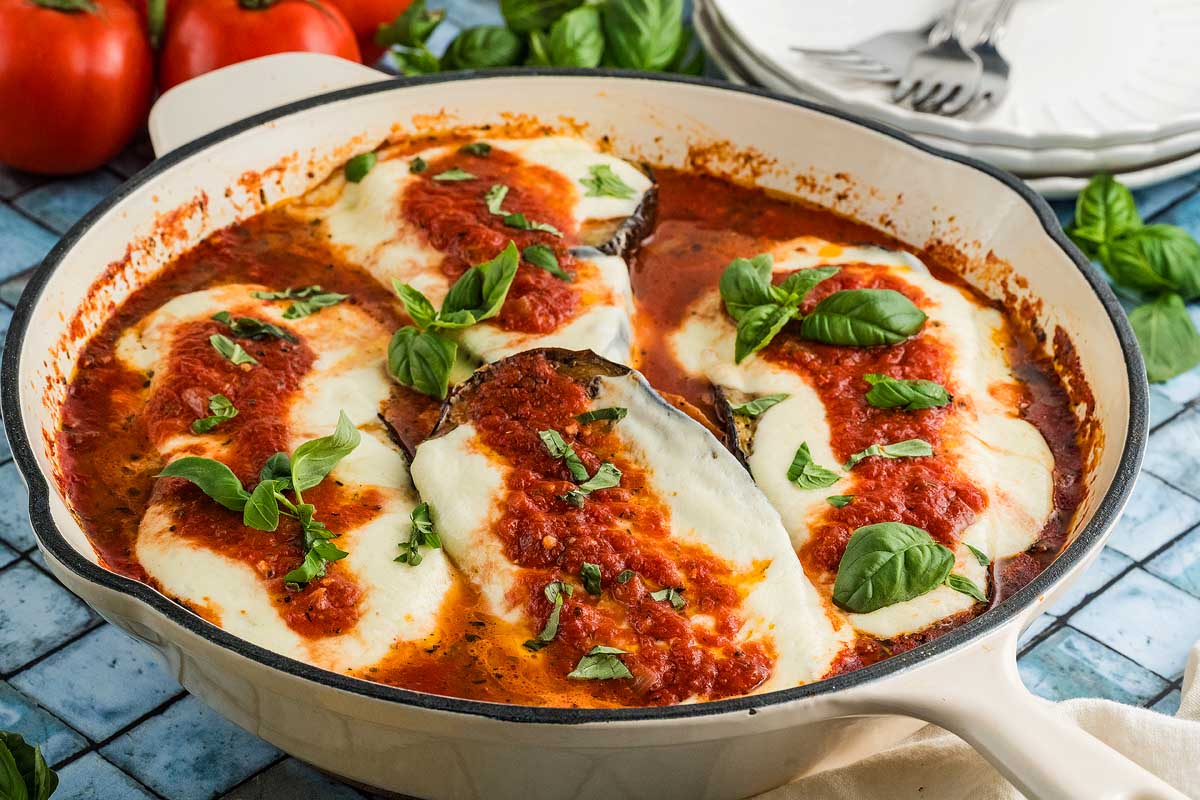 A skillet with baked eggplant slices topped with melted cheese, tomato sauce, and fresh basil leaves, with plates and tomatoes in the background.