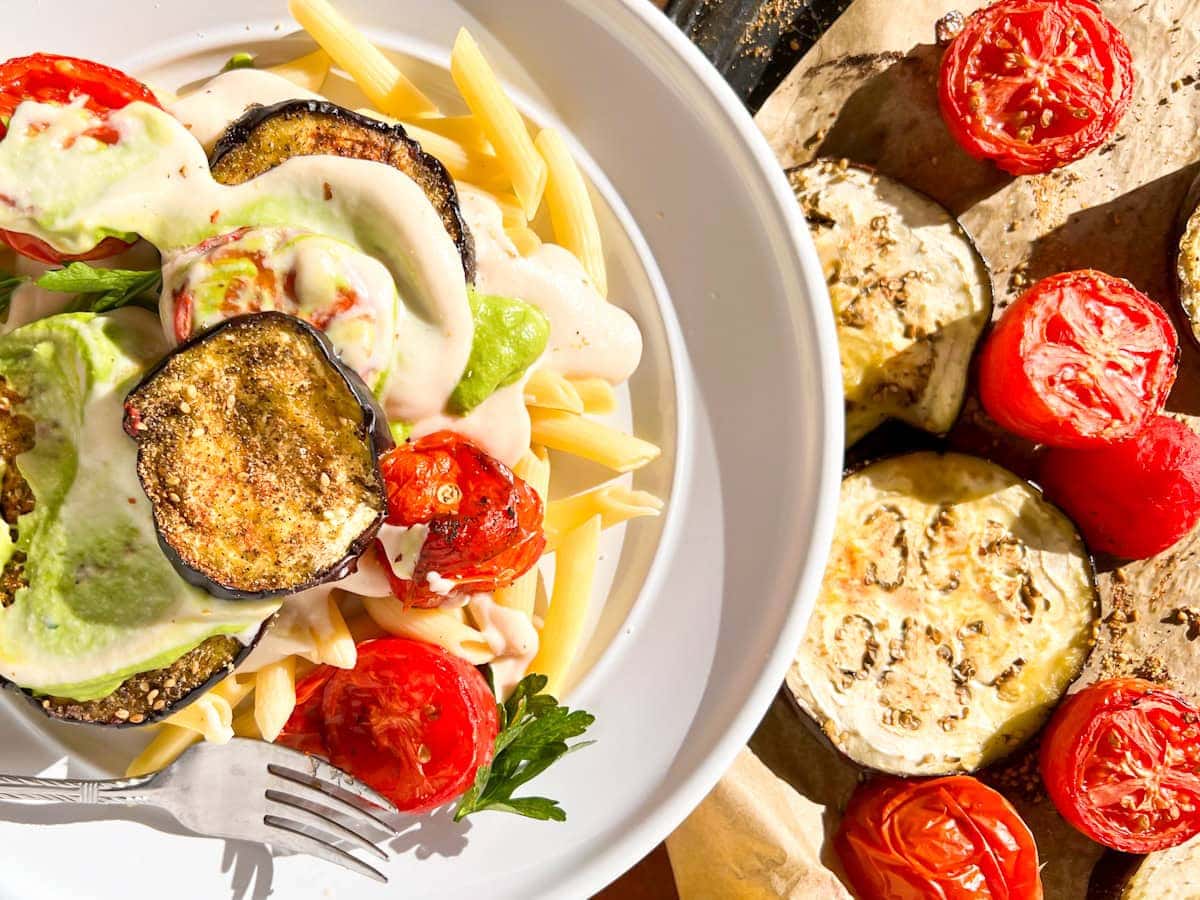 A plate of penne pasta topped with roasted eggplant, tomatoes, and creamy sauce, next to a tray of roasted eggplant and tomatoes.