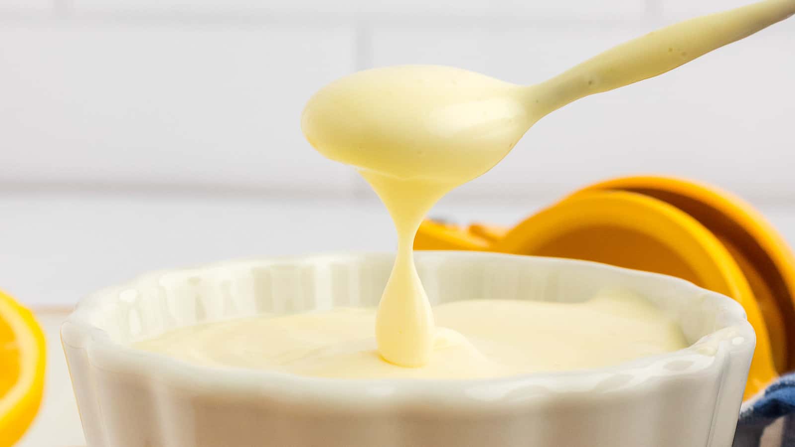 A spoon lifts thick, creamy sauce from a white ramekin, with a yellow citrus juicer visible in the background.