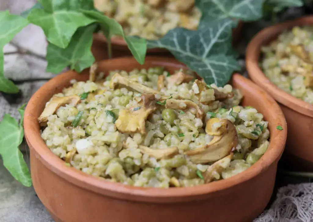 A clay bowl filled with rice, green peas, and sliced mushrooms, garnished with herbs, with green leaves in the background.
