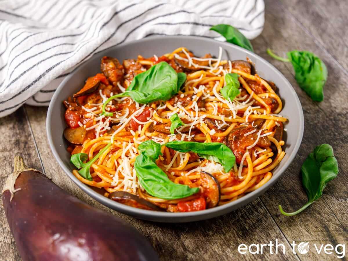 A bowl of spaghetti with tomato sauce, sliced eggplant, grated cheese, and fresh basil leaves, placed on a wooden table with a striped cloth and an eggplant nearby.