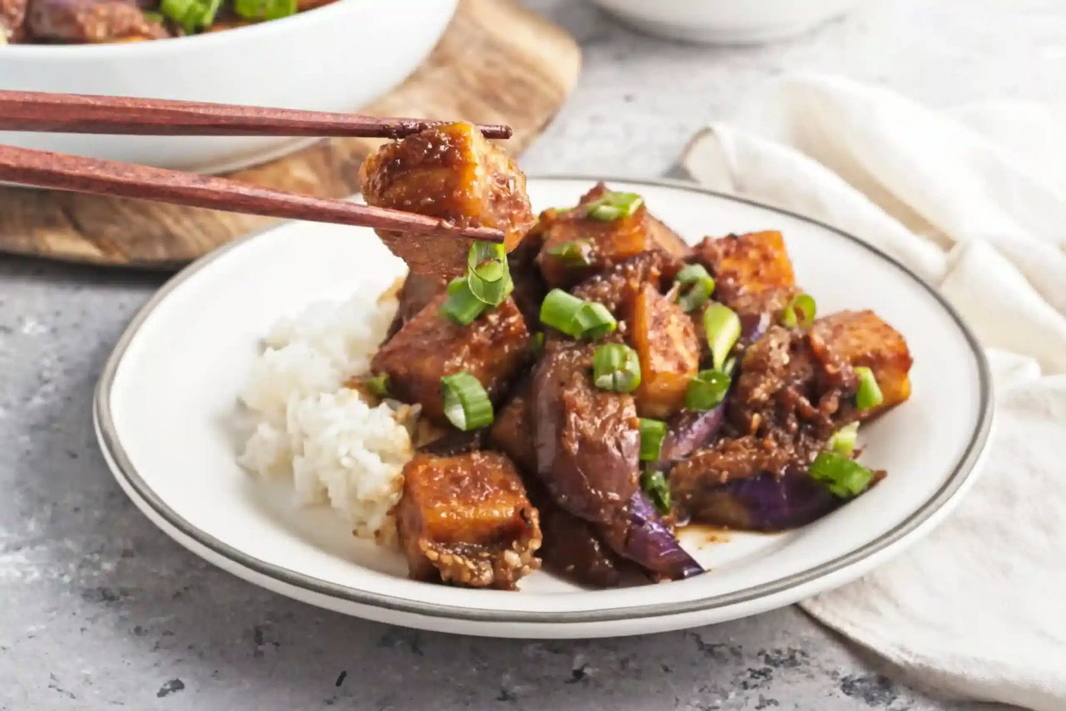 A plate of white rice topped with stir-fried tofu, eggplant, and green onions, with chopsticks holding a piece of tofu.