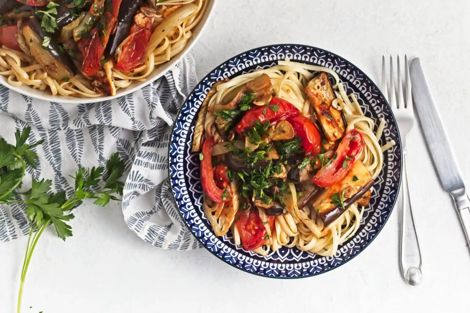 A bowl of pasta topped with roasted vegetables and herbs sits next to a partial view of another bowl on a patterned cloth, with a fork, knife, and parsley nearby.
